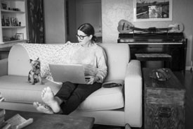 A young woman sitting on her couch with her dog using her laptop researching if she should buy a house or an apartment.
