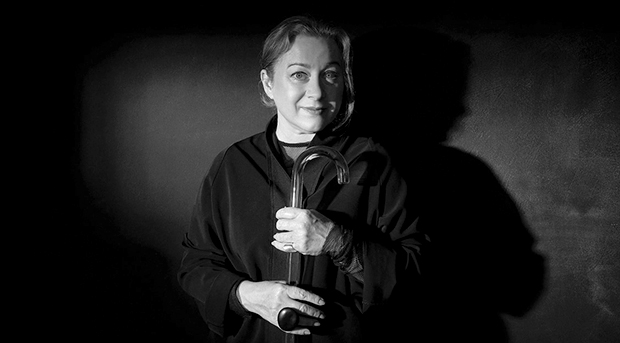 A greyscale portrait of Gill Hicks standing against a blank wall and looking at the camera with a closed-mouth smile. She is holding a walking stick to her torso.