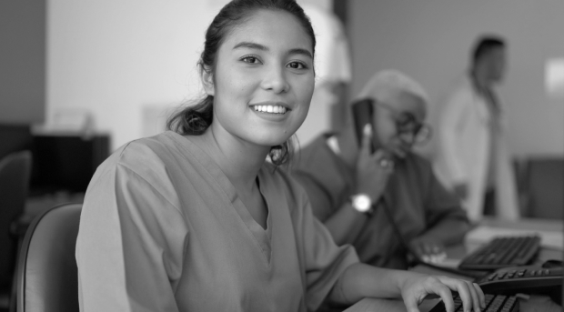 A health professional wearing scrubs is sitting at a desk and smiling at the camera. A woman behind her is on the phone and a doctor in a lab coat is passing in the background.