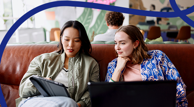 Two women sitting on a lounge looking at a tablet computer and laptop.