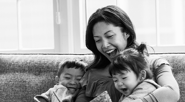 A mother sits on a couch holding her two toddler children and reading to them from a book. All three are laughing.