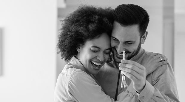 A young man and woman hold each other in a living room and smile happily. The man is holding the keys to their first home.
