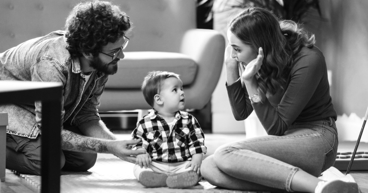 A man and a woman play with their baby on the floor of the living room in their home.