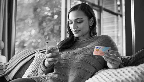 A woman is sitting on a couch using a mobile phone and holding a Health Professionals Bank Credit card. In the background is a window to the garden.