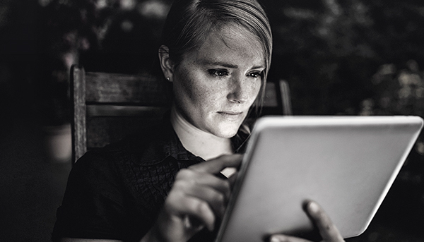 A close up image of a serious-looking woman using a tablet device while sitting in a chair. The background is dark.