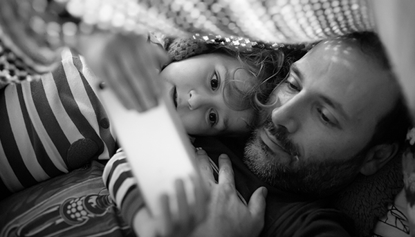 A greyscale close up image of a father and child looking at a mobile phone. A blanket is draped over them.