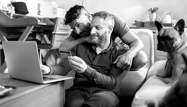 A man holding a bank card sits on the living room floor with his partner leaning on his shoulder. Both are looking at a laptop in front of them. A French bulldog sits on a couch to their right.