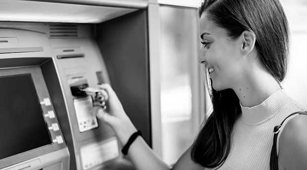 A young woman is smiling as she inserts her bank card into an ATM.