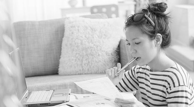 A woman sits in thought with a pen in her mouth, her laptop and financial documents, working out her budget.