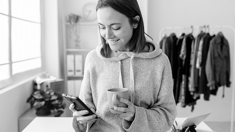 A woman looking at a phone in her right hand while holding a mug in her other hand.