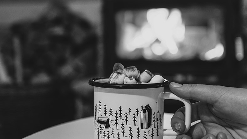 A hand clutches a mug of hot chocolate in front of a roaring fireplace.