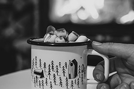 A hand clutches a mug of hot chocolate in front of a roaring fireplace.