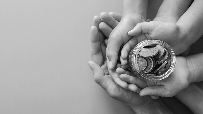 Two pairs of hands, one a child's and one an adult's, clasp a full jar of money.