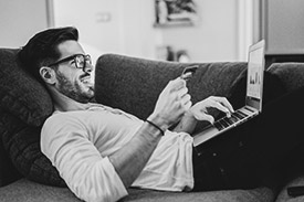 Man wearing glasses laying on a couch with his credit card and laptop.