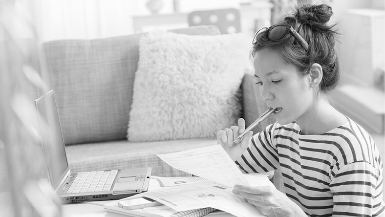 A woman sits in thought with a pen in her mouth, her laptop and financial documents, working out her budget.