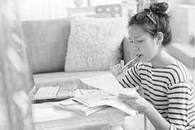 A woman sits in thought with a pen in her mouth, her laptop and financial documents, working out her budget.