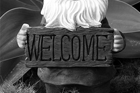 A happy garden gnome sits next to a garden plant holding a sign that reads welcome.