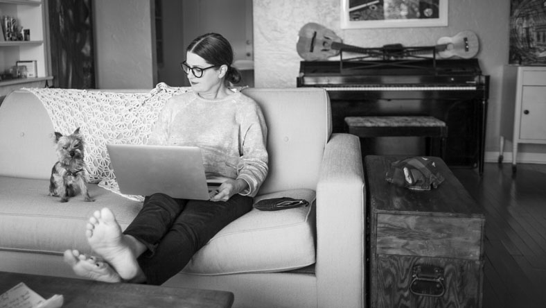 A young woman sitting on her couch with her dog using her laptop researching if she should buy a house or an apartment.