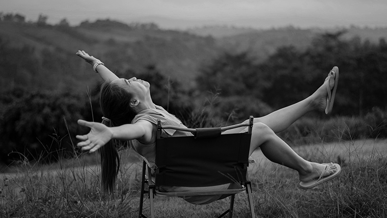 A black and white photo of a woman relaxing in a chair in an open field.