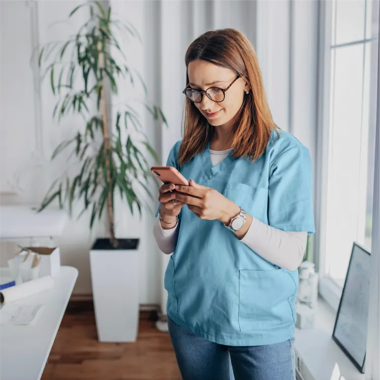 A healthcare professional in light blue scrubs stands indoors using a smartphone