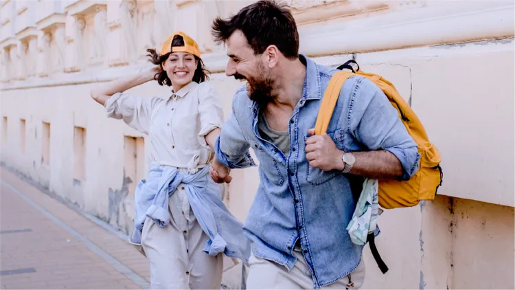 Young couple laughing with the male carrying a yellow backpack and the female wearing a yellow hat.