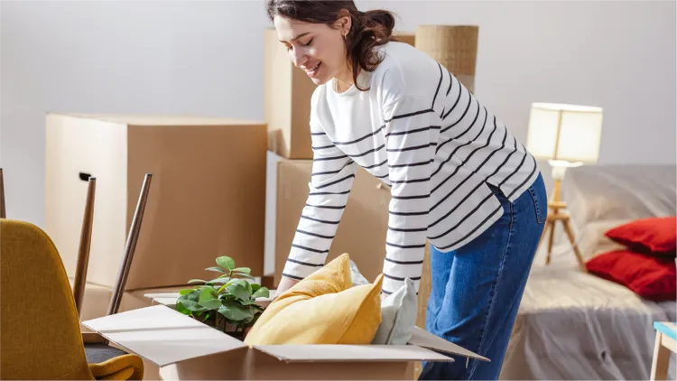 A woman is packing household items into a cardboard box in a room filled with moving boxes.