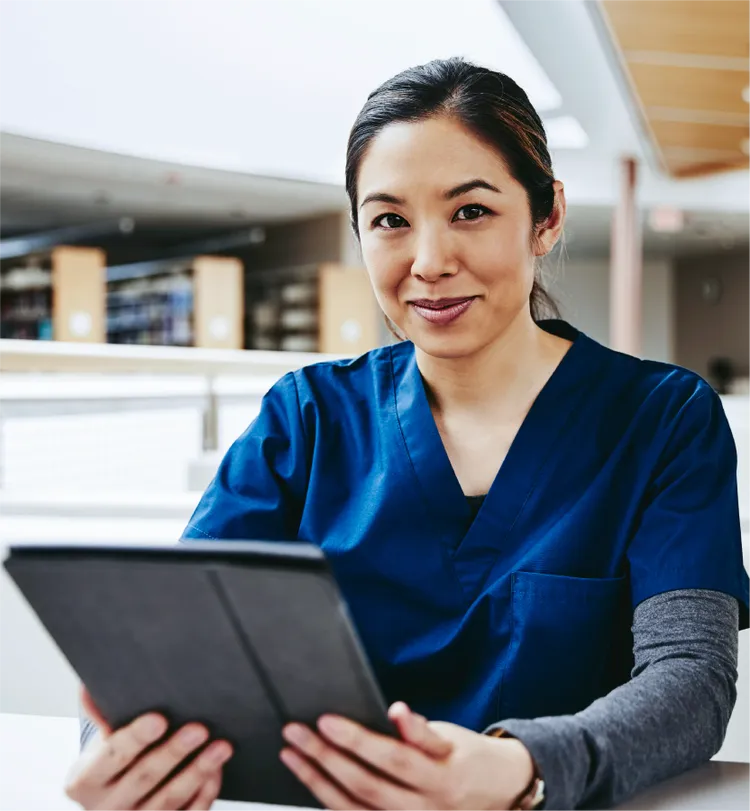 A healthcare professional in blue scrubs sits indoors holding a digital tablet.