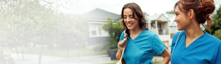 Two healthcare workers in blue scrubs walk side by side outdoors in a residential neighbourhood.