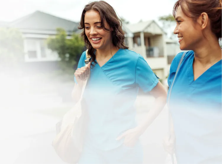 Two healthcare workers in blue scrubs walk side by side outdoors in a residential neighbourhood.